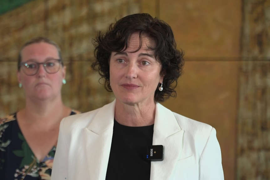 A woman with short brown hair and a white blazer faces the camera. 