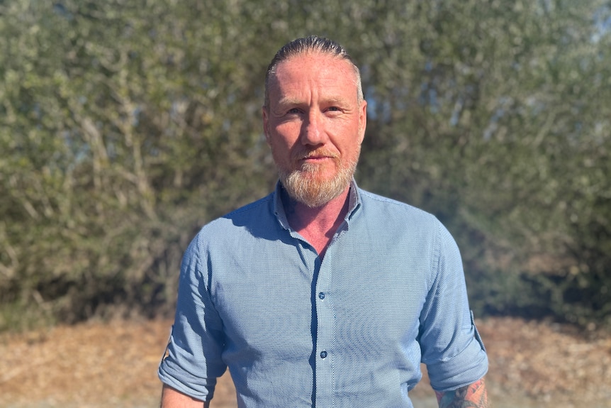 A man with grey hair and a beard in a blue button up shirt.