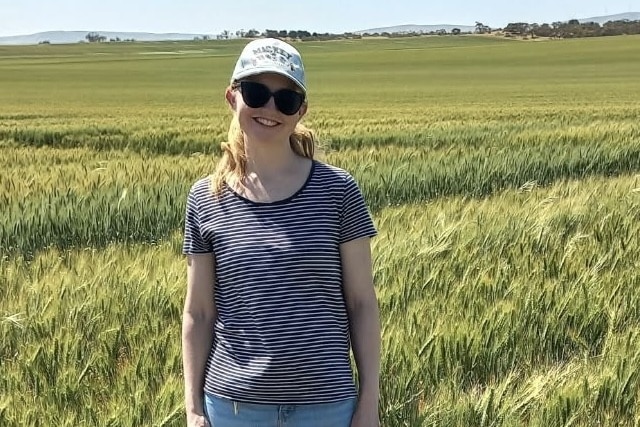 A smiling woman in a cap and t shirt stands in a field of wheat.