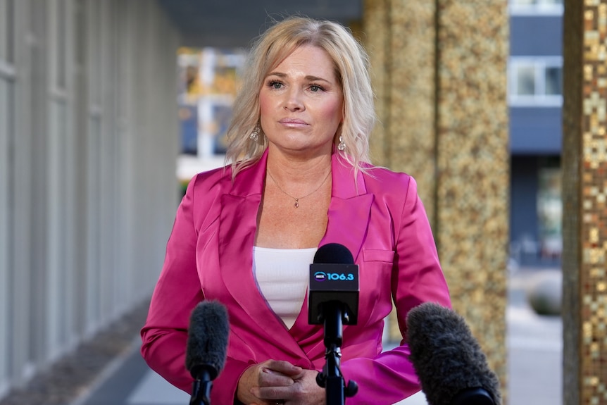 A politician in a pink jacket stands in front of microphones.