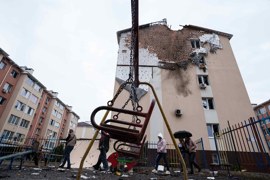 A damaged apartment building sits behind a rusty-looking metal swingset against a grey sky.
