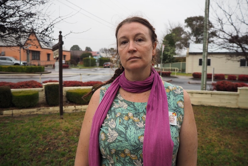 Woman wearing scarf and floral dress looks into camera. Behind her is trees and houses.