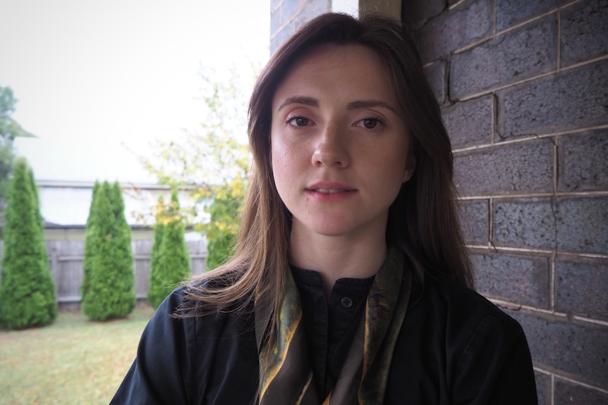 Woman standing next to brick wall looks directly into camera with neutral expression. behind her are trees. 