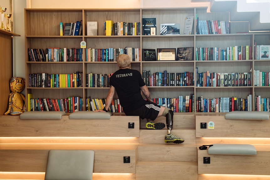 Man with amputated legs and prosthetics looks at library, his shirt reads 'veterans'. 