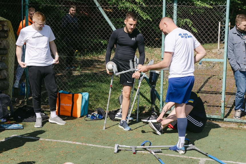 Two men, each with an amputated leg stand on a football field, beside them are two other men watching on. 