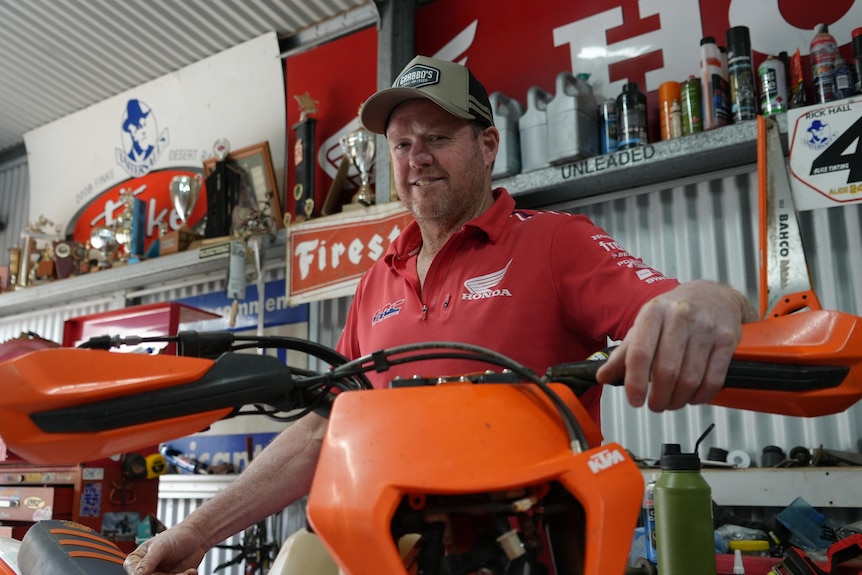The camera looks up and through the handlebars of an orange motorbike to a smiling man in a red honda shirt in a shed.