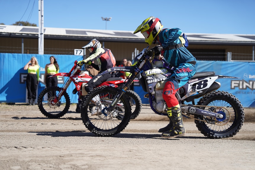 Wide shot of two motorcyclists at the start line, dirt road, blue banners in the background.