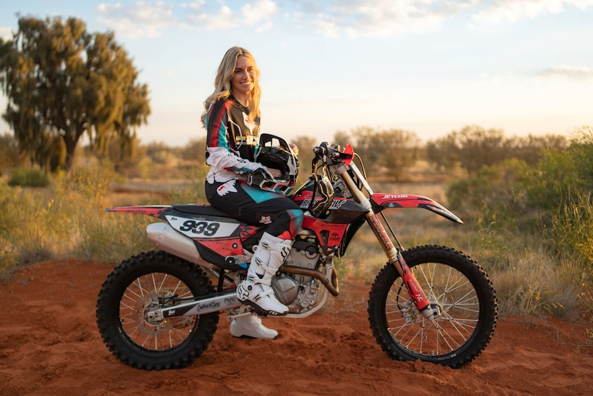 Blonde-haired woman sitting on her motocross bike, with the Central Australian desert as her backdrop