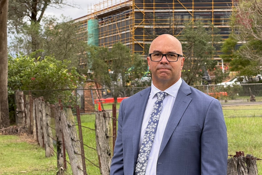 Andrew Carfield, wearing glasses, a suit, stands in front of a green area and construction apartment site.