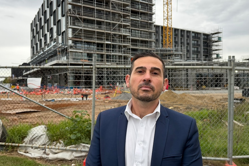 Adam Leto in a navy suit, stands in front of an apartment construction site.