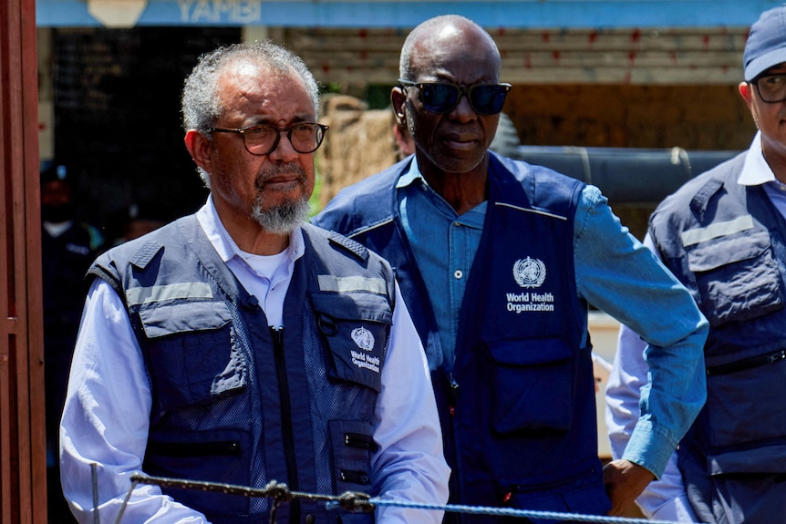 Three men standing wearing navy blue vests and blue shirts.