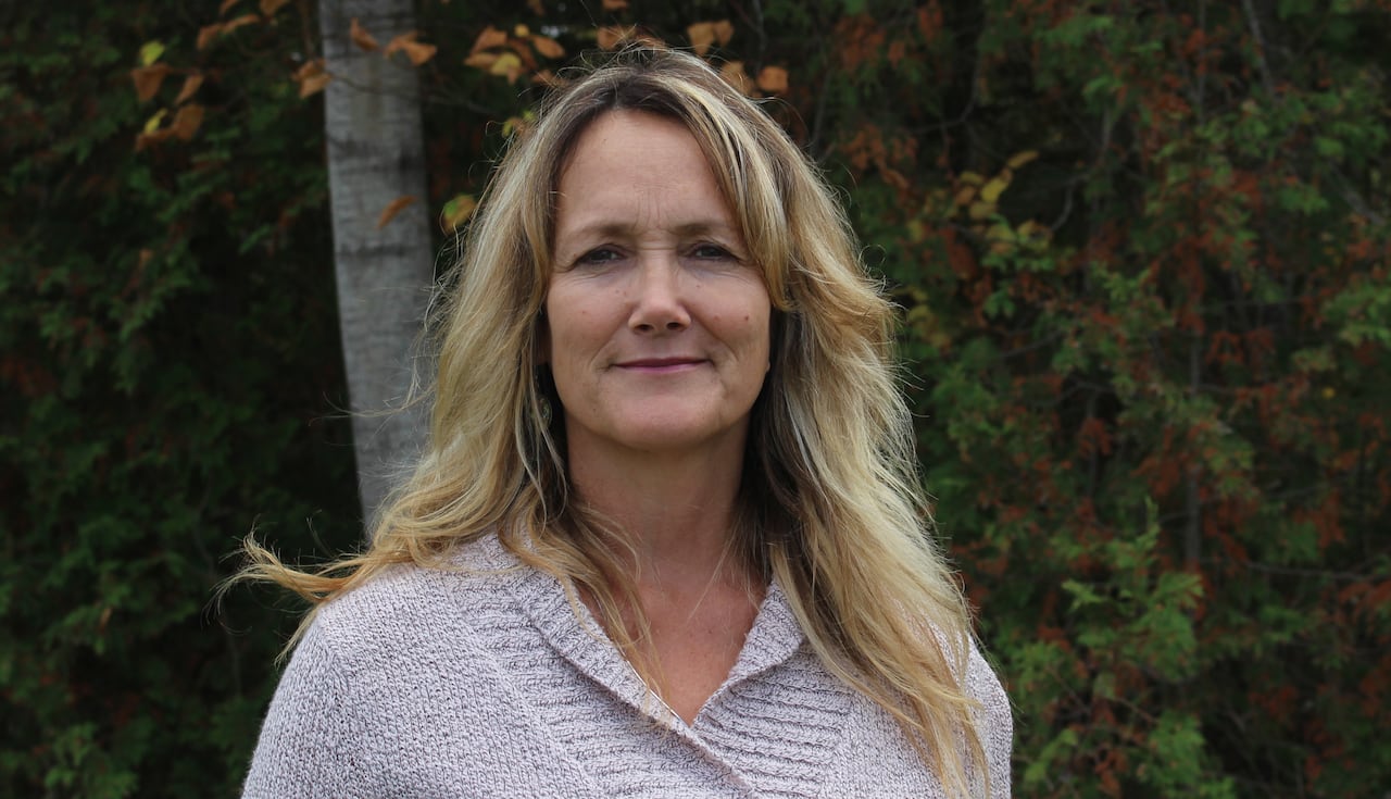 A woman wearing a sweaters stands outdoors with trees and remaining fall foliage behind her. 