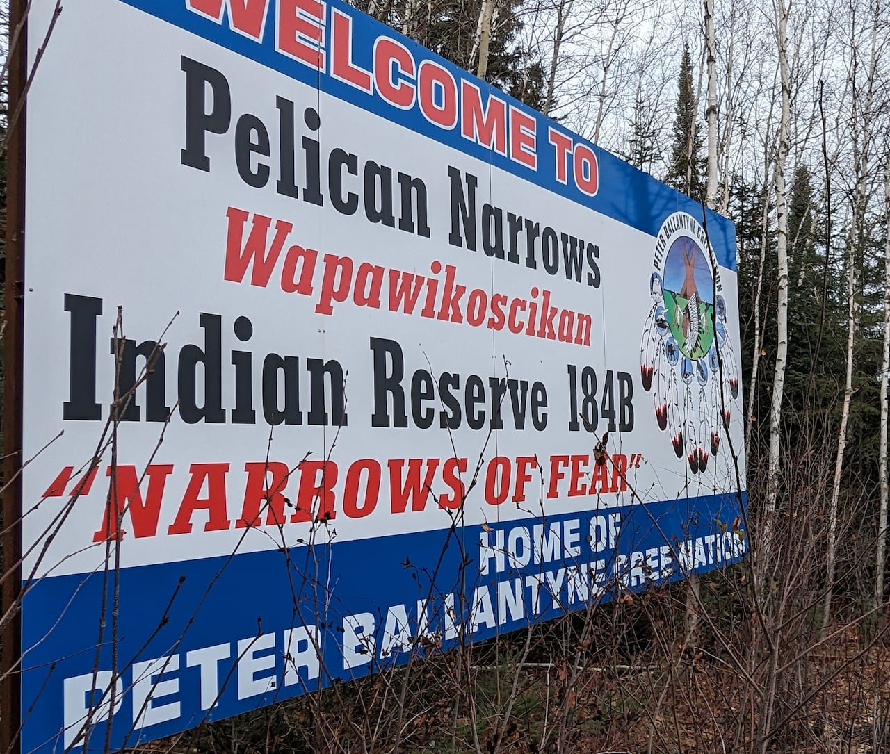A Pelican Narrows welcome sign overlooking a bridge, with a red dress hung on a nearby tree.