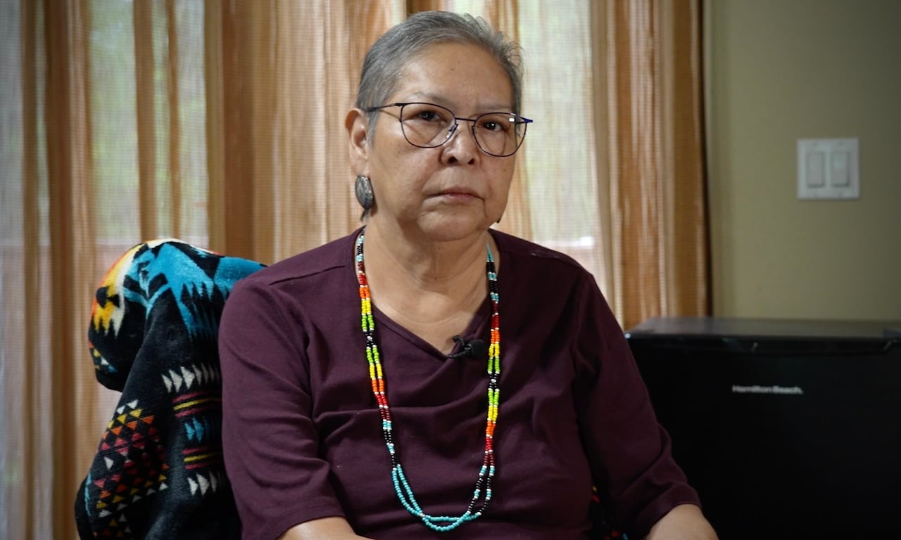 An elderly, tan, seated woman looks into the camera wearing a purple shirt.