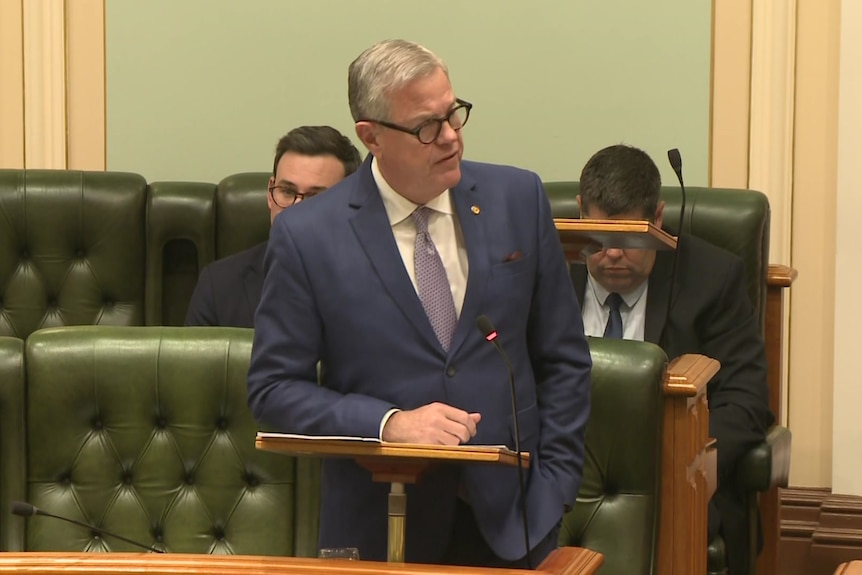 A man in a suit and glasses speaking in parliament. 