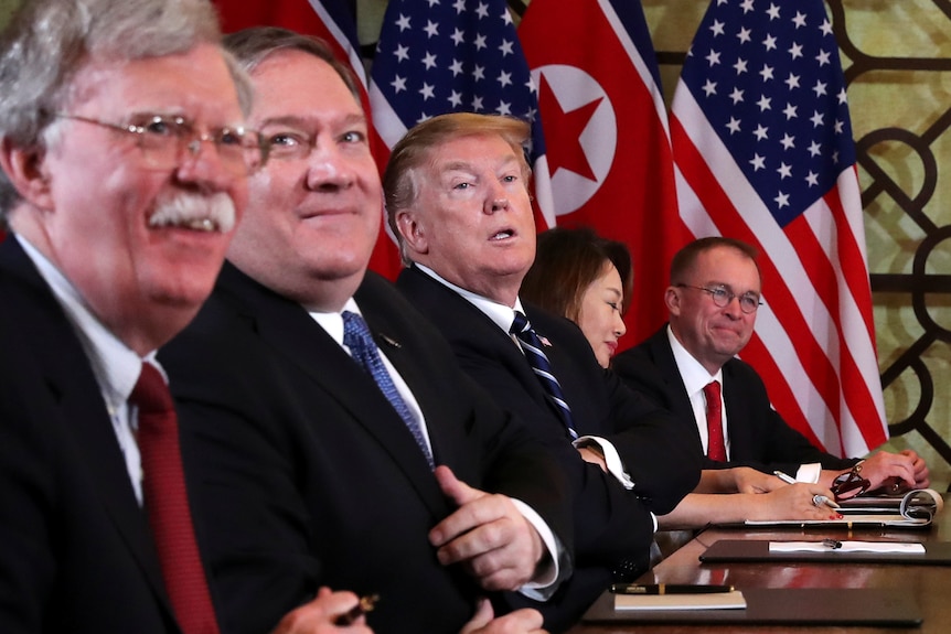 John Bolton, Mike Pompeo and Donald Trump sitting at a table with North Korean and US flags behind them.