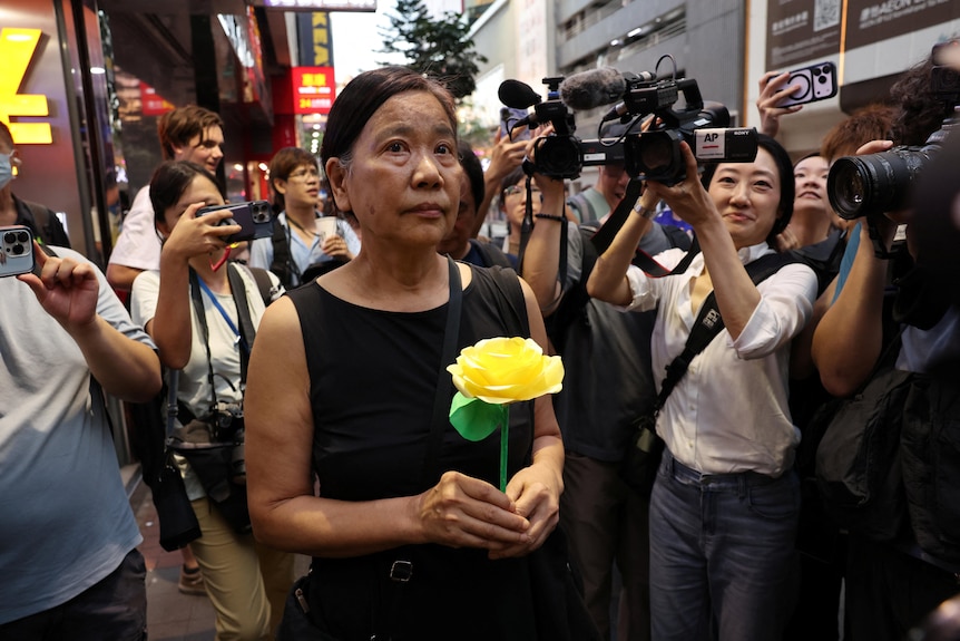 A woman holds a yellow rose and is surrounded by photographers.