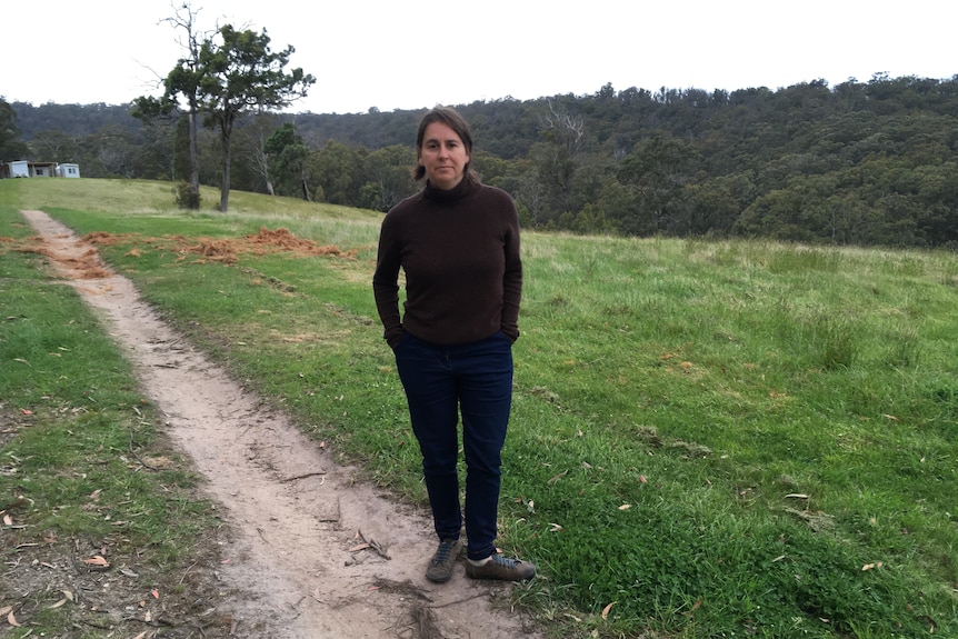 Woman wearing warm clothes stands on a track surrounded by grass, with trees and a building in the background.