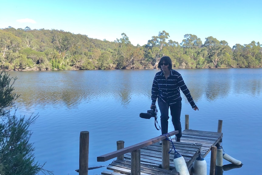 Woman holding a small camera in her hand walks up a small wooden jetty on a river.
