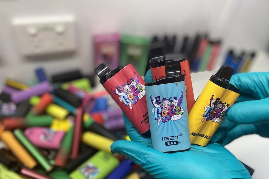 Gloved hands hold a selection of vapes above a bin of multi-coloured objects.
