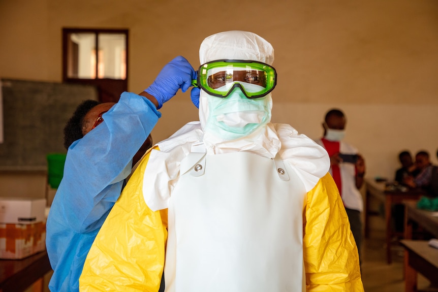 A person in scrubs applies a mask to a person in full PPE.