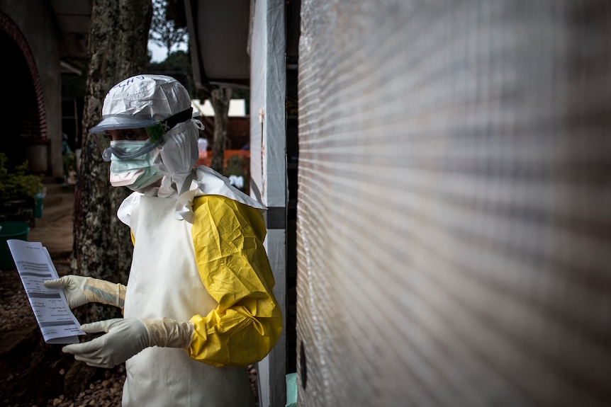 A woman in PPE stands holding a flyer outside a building.