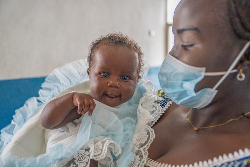 A baby held by its mother who wears a face mask reaches towards the camera.