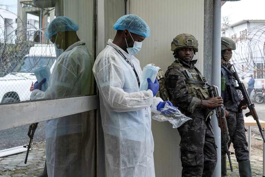A doctor in a lab coat and mask stands next to two military men holding rifles.