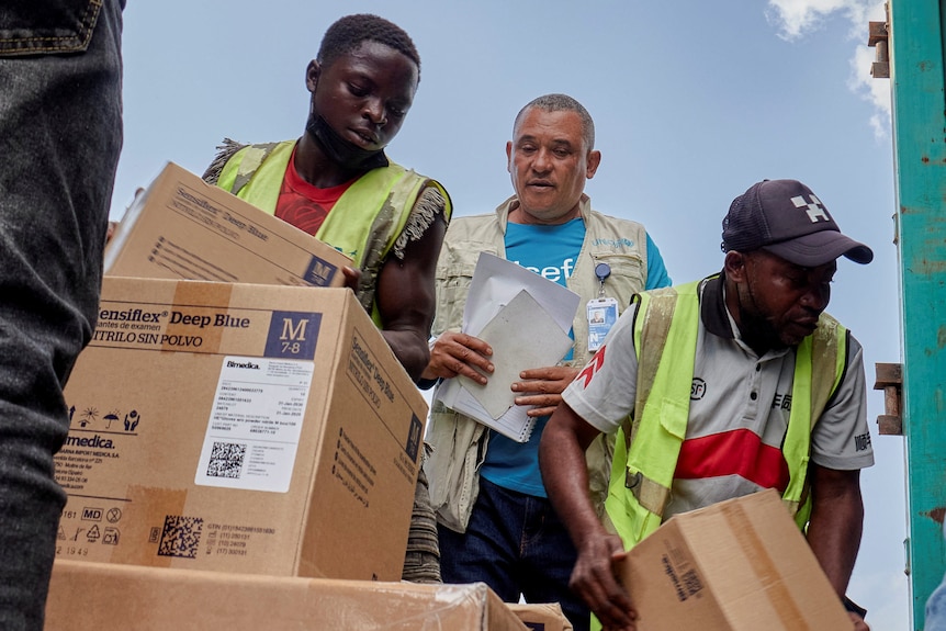 staff in high vis lift boxes as a man in a blue shirt watches them 