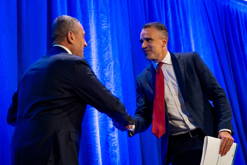 Two men wearing suits stand on stage looking at each other, smiling and shaking hands. 