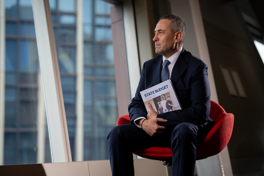 A man wearing a navy suit and blue tie sits in a red chair in front of a window holding a booklet that says State Budget 2026-27