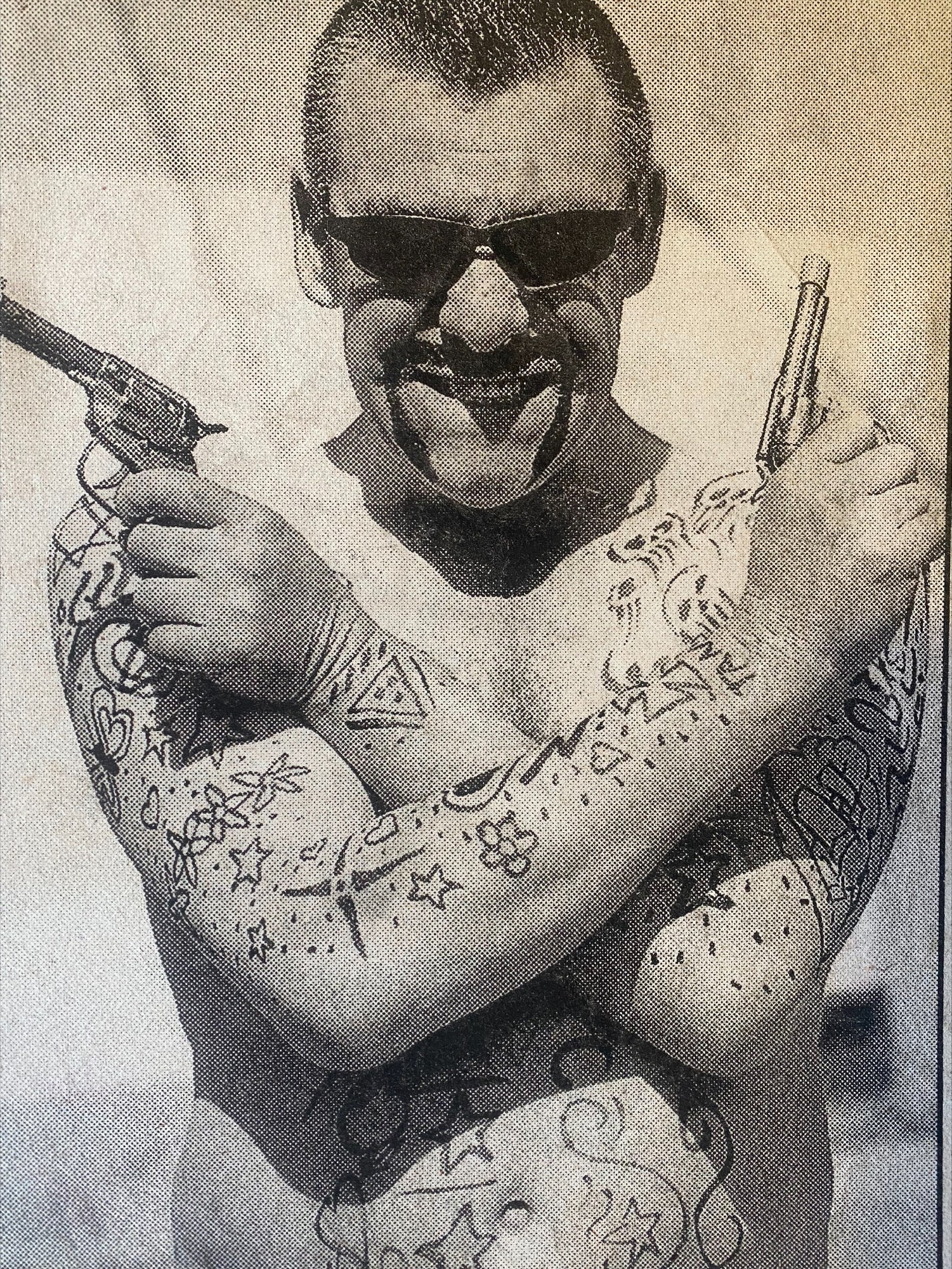 A black and white photo of a shirtless man with hand drawn tattoos, sunglasses and fake guns