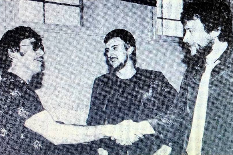 A black and white image of two men shaking hands and another man in the background over text