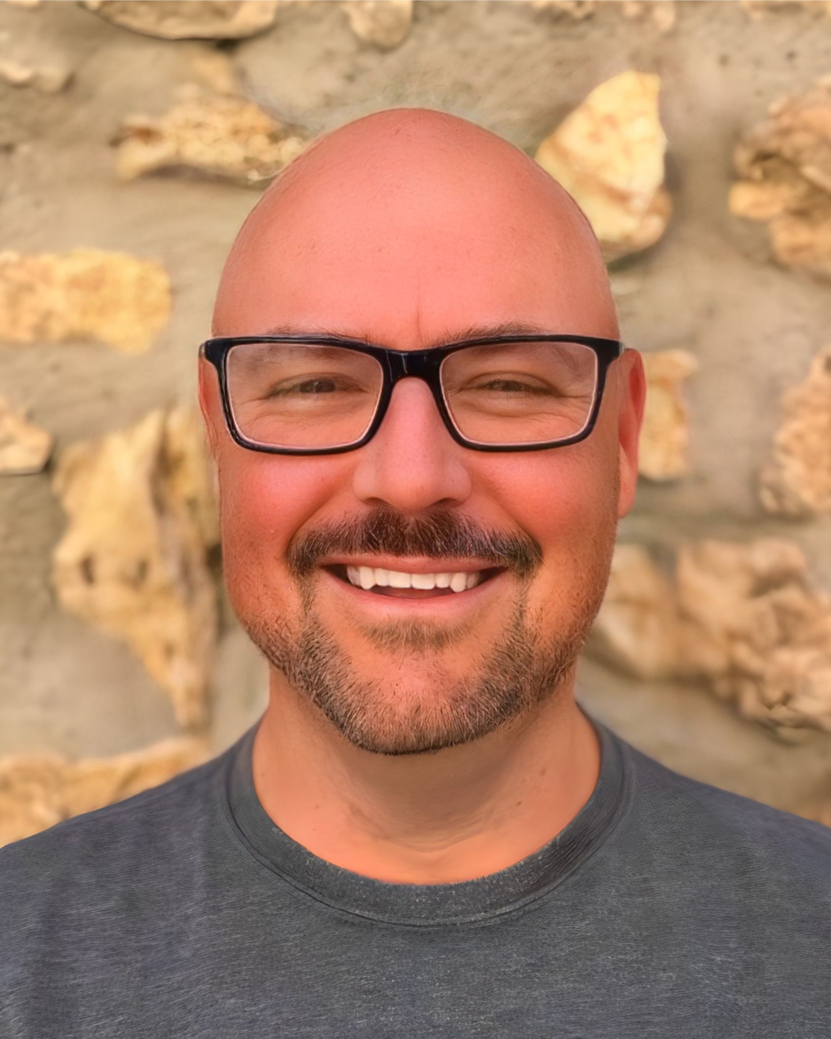 A bald man wearing black rimmed glasses standing in front of a wall and smiling at the camera 