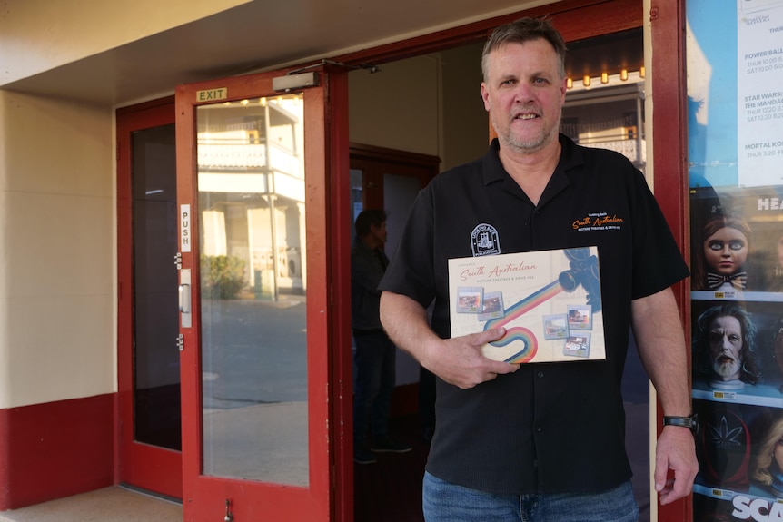 A man standing outside a cinema building and holding a book on South Australian cinemas