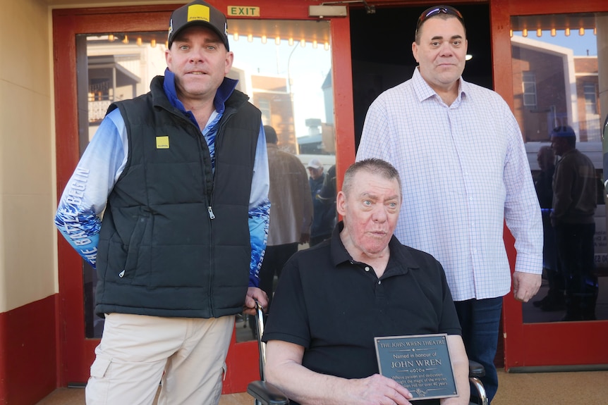 Two men standing on either side of an older man in a wheelchair who is holding a plaque outside a cinema