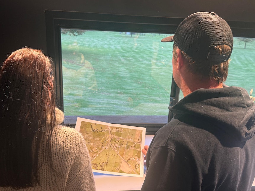 A photo of a man and woman, standing in a kitchen holding a map, looking out the kitchen window into a peaceful backyard