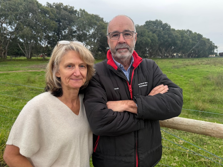 A photo of a blonde woman wearing a white top, standing next to a man with glasses wearing a black jacket, standing at a farm