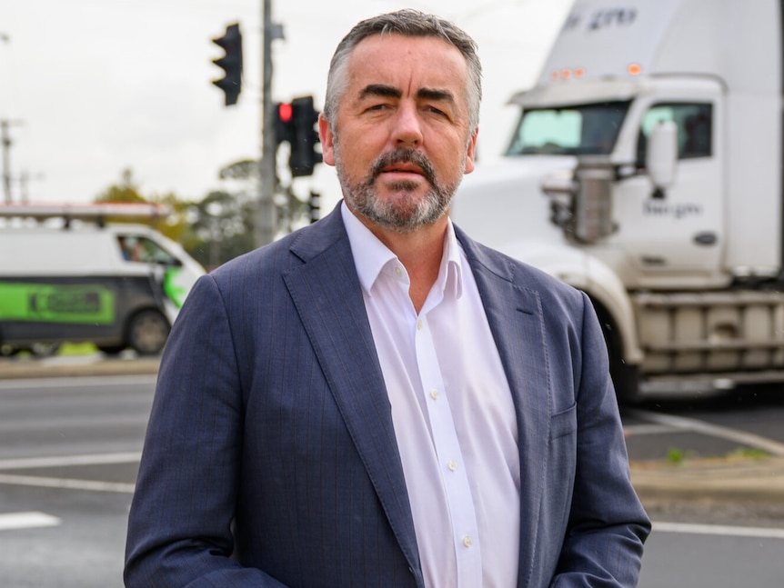 A photo of a man wearing a white shirt and blue blazer, standing at an intersection, with a truck and van in the background