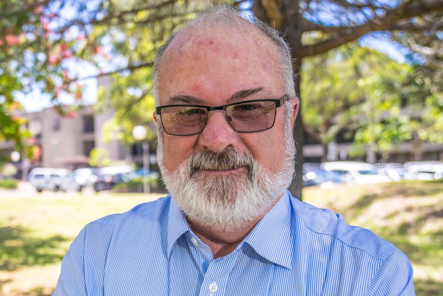 A photo of a man with a grey beard, wearing glasses and a blue buttoned shirt, crossing his arms, standing outside