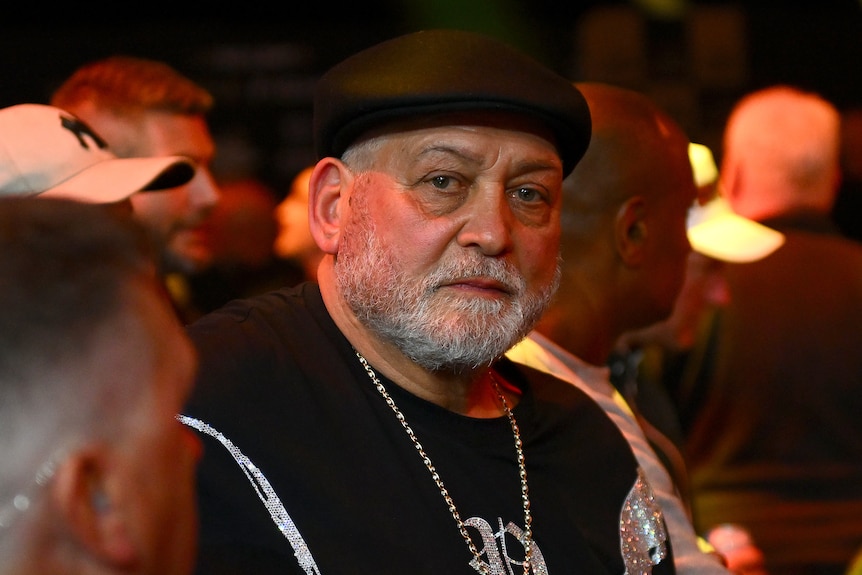 A man wearing a hat and bejewelled chain sits among a crowd