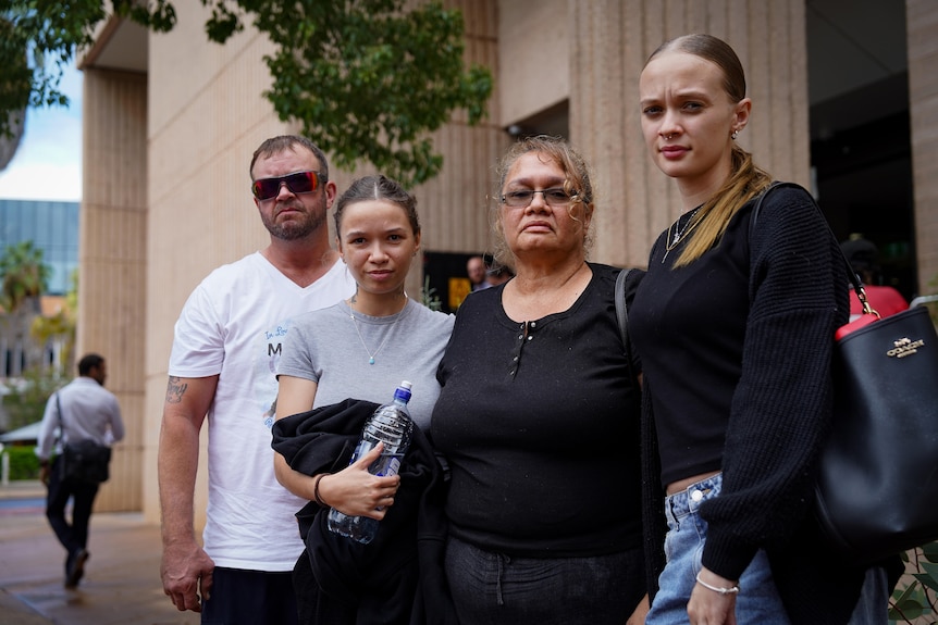 Four people, looking serious, standing outside a court house. 