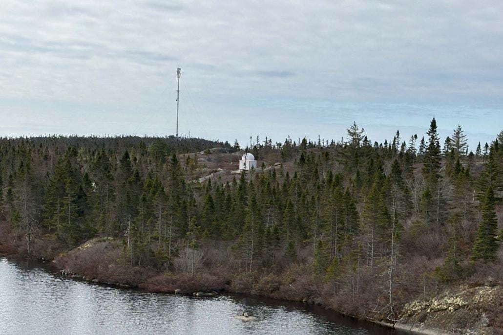 A white domed structure sits on a rocky ridge near a lake's edge, in a cleared space. Trees and shrubs are in a wooded area nearby