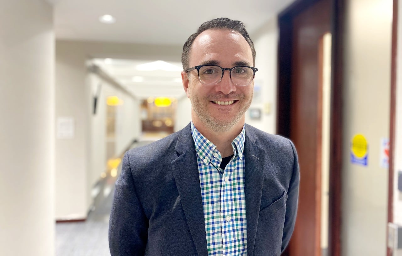 A white man with short dark hair and glasses smiles at the camera as he stands in a hallway. He wears a checkered shirt and navy blazer.