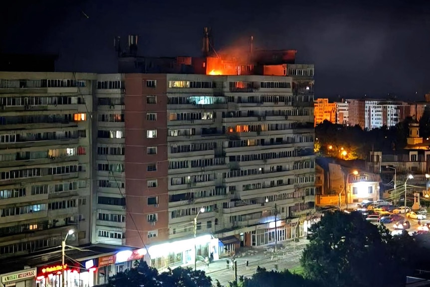 an apartment block at night, the roof is on fire after being struck