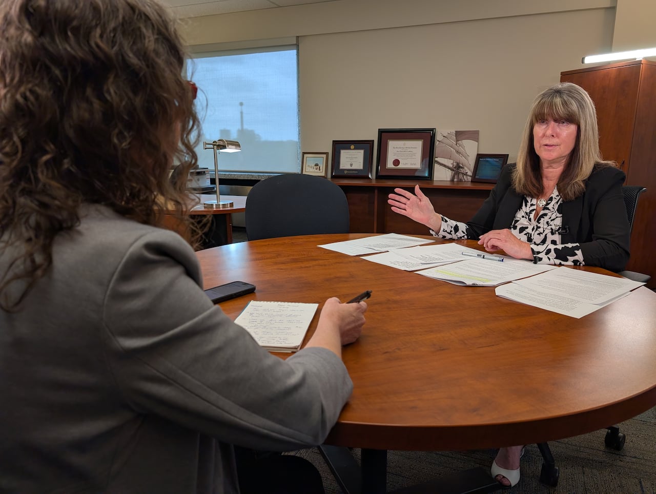 A woman with straight blond hair sits at a round table with several papers spread in front of her. She sits across from a reporter with curly brown hair.