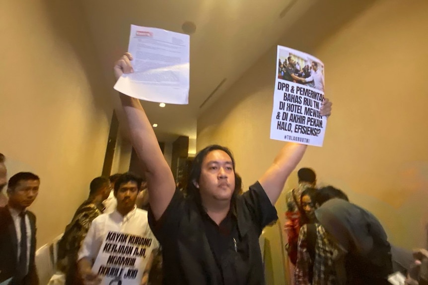 A man with shoulder-length hair, dressed in black, raises two posters, one in each hand.