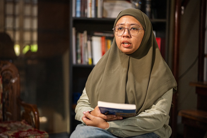 A woman wearing glasses and a hijab holds a book while sitting in a chair.