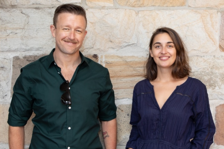 Man and woman stand next to each other in front of a stone wall, smiling.