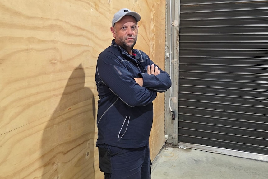 Man wearing blue jumper and shorts and a grey cap stands in a shed with his arms crossed, looking disappointed.